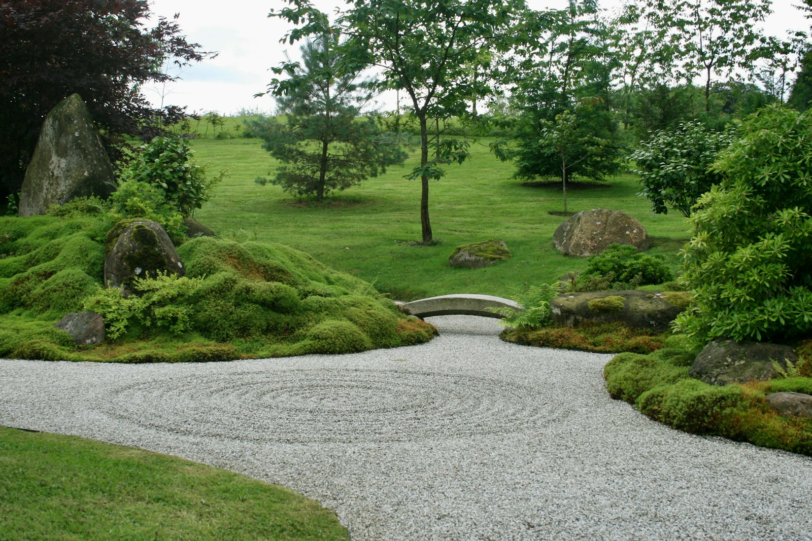 Zen garden featuring raked gravel forming concentric circles, surrounded by lush green moss, rocks, and trees, creating a serene landscape.